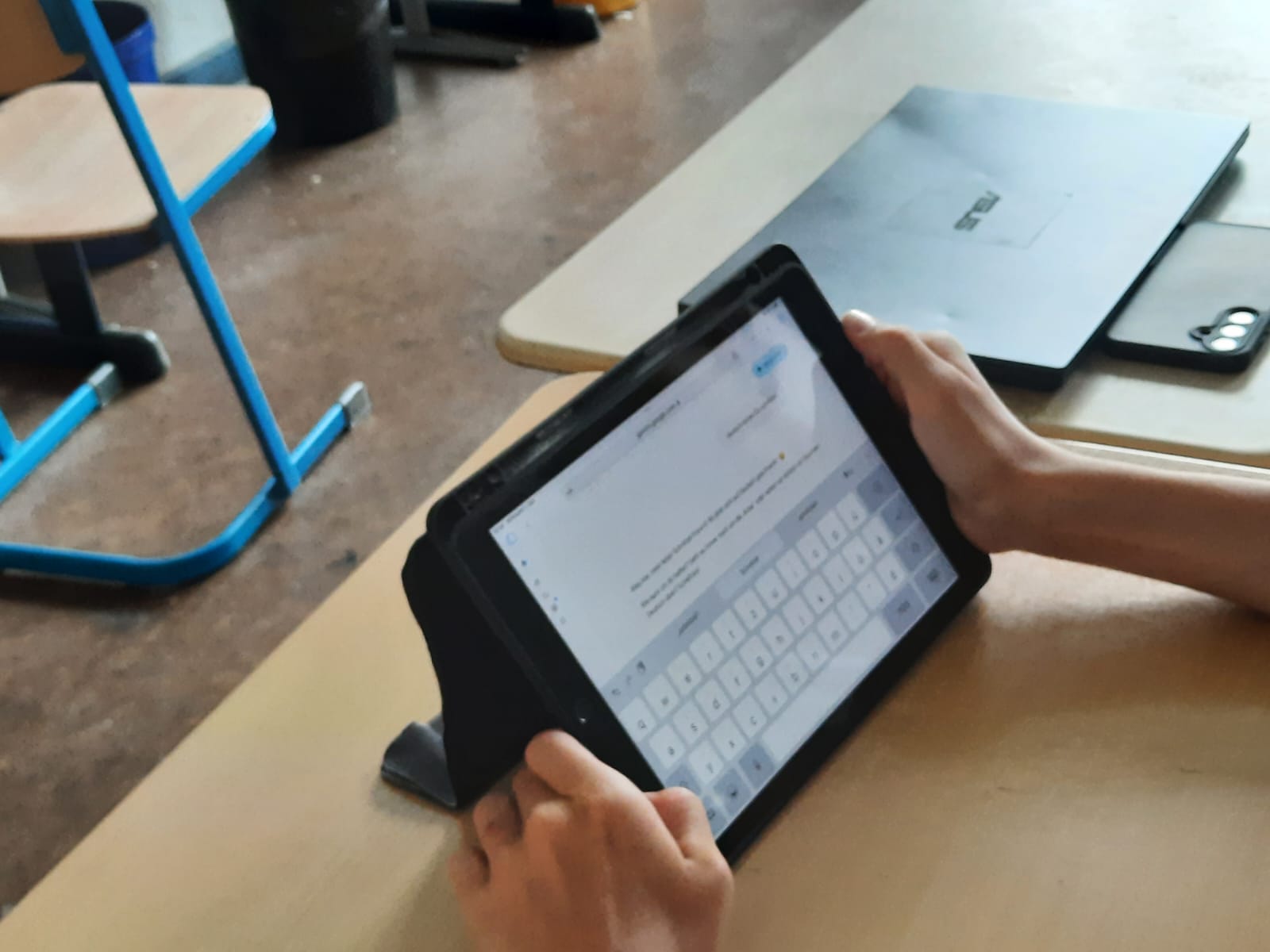 Schüler tippt auf einem Tablet in einem Chat-Fenster, im Hintergrund liegen Laptop und Smartphone auf dem Tisch